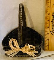 Front view of dark woven basket with braided handle and raffia bow alongside ruler for scale