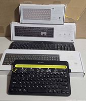 Stacked view of three boxed Vital keyboards, one unboxed black keyboard, and Logitech K480 in front