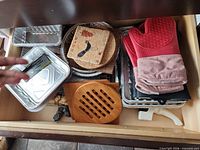 Drawer view showing aluminum pans, oven mitts, cork trivets, wood trivet, knife sharpener, partial elephant holder