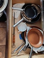 Drawer view showing stacked black and copper frying pans, sharpening steel, cup and protectors