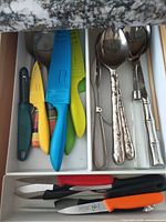 Drawer view showing colourful sheathed paring knives, hammered-handle spoons, bamboo-style spoon, peeler, and additional knives