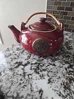 Side view of red glazed stoneware teapot with bamboo handle and brass-tone medallion