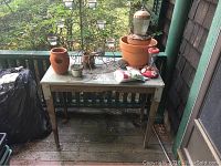 Wood and metal potting bench on wooden porch floor with various pots and candleholders placed on top.