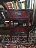Front view of the vintage mahogany side table showing drawer, legs, lower shelf, and overall shape. Backdrop includes books on a shelf.