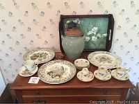 Full view of decorative china and serving items arranged on wooden table including plates with bird and floral motifs, cups and saucers, serving urn with ladle and wooden tray with floral painting.