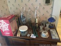 Photo showing assortment of candleholders, hurricanes, lantern, candles, and decorative items on a wooden surface against floral wallpaper.