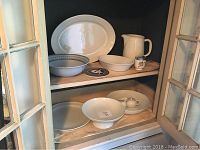 Collection of 9 pieces of tableware displayed on shelves in a cabinet including plates, bowls, pitcher, creamer, sugar bowl, and decorative item.