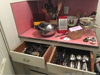 Overview of countertop with nested stainless steel bowls, grater, metal food mill, and drawers opened showing various forks, knives, and spoons.