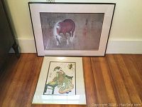 Image of both framed prints showing the horse print and the woman print lying on a wooden floor and leaning against a white wall.