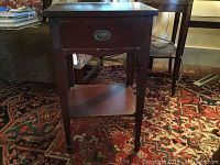 Front view of the small mahogany finished side table showing the single drawer with oval metal pull missing the handle and the lower shelf.
