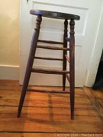 Front angle view of antique tall stool showing round oak seat and turned wood legs with three rungs.