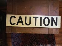 Full view of the metal caution sign laid flat on a wooden surface, showing the entire word CAUTION in black letters on a white or light beige background.