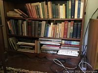 Photo showing multiple wooden shelves filled with hardcover and paperback books in varying conditions. Books are visibly dusty and some stacked horizontally.