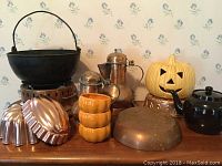 Overall view of 13 items on table including cast iron pot, copper pitchers, ceramic pumpkin, aluminum molds, teapots, and bowls