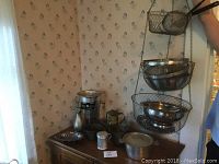 Overview of metal kitchen items arranged on wooden cabinet including a champagne bucket, hanging wire basket, several metal bowls and colanders