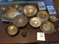 Photo showing vintage silver plated tableware arranged on table including large scalloped platter, pedestal compote, round plates and bowls, small ladle, and other pieces with heavy tarnish.