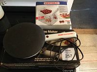 Maxim electric crepe maker showing round black cooking surface and power cord with light indicators on handle, alongside Leifheit cherry pitter box.