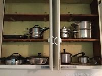 Kitchen cabinet shelves containing assortment of used kitchen pans including double boilers, covered fry pans, skillets, and saucepans in different sizes, mostly stainless steel with black handles and knobs.