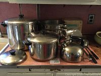 Two large Revere stainless steel stockpots with lids alongside a shiny Revere tea kettle, a KitchenAid food chopper attachment, an anonymous stainless steel pasta pan, and three kitchen utensils including knives and a peeler, all on a kitchen counter.