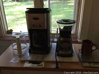 Full coffee station setup with coffee maker, grinder, scale with mug, and marble slab base shown on wooden table in front of window.