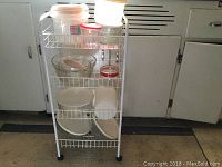 Full view of the white wire utility cart holding various glass and plastic kitchen storage containers arranged on all shelves.