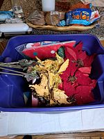 Blue plastic tote containing artificial red poinsettias, gold stems, holiday tin and packaged items