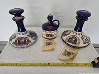 All three ceramic rum decanters with booklets and tape measure for scale