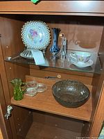 Overview of cabinet shelf showing plate, dome lid, blue vase, clear stands, teacup and saucer, lower shelf items