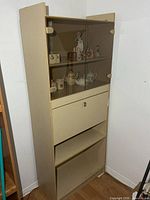 Full front view of beige display cabinet showing upper glass doors, drop-front desk, and lower shelves