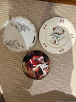 Three plates laid out: floral handled cake plate, Canada–US commemorative plate, Santa Claus Christmas plate