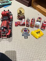 Group shot showing all items laid out, including large metal truck, snow globe, eight small vehicles, IAFF emblem, metal tag, yellow plastic items