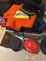 Box showing orange jacket, peaked cap, navy garments, additional caps, red helmet