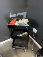 Front angled view of black 3-tier cart on wheels with top tray holding assorted rollers and clips