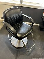 Front view of black vinyl salon chair showing seat, armrests, footrest and round hydraulic base