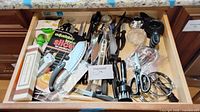 Drawer view displaying majority of utensils including knives, shears, measuring cups, peeler, scoop, tongs