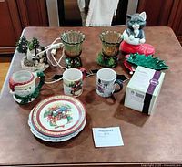Overview of lot on table showing plates, mugs, holly dish, candle holders, cat figurine, Scentsy box, tree decor