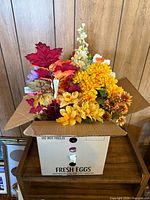 Open cardboard box labeled Fresh Eggs filled with varied artificial flowers and foliage stems in autumn colors