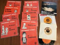 Photo showing 9 Herbert Hisel 45 RPM records in red illustrated sleeves and 3 other 45s including one Mac Davis and two others with various covers