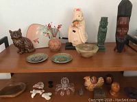 Photo of assorted decorative items on wooden shelf including brown ceramic cat, painted eggs in rooster-shaped container, clear glass octopus figurine, glass bowl, pottery bowl, carved wooden statues, and small animal figurines.