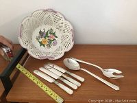 Overall view of the porcelain serving bowl, four knives, and two salad servers on a wooden surface with measuring tape for scale.