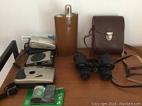 Photo shows three vintage Instamatic cameras, Ralik 8x30 binoculars with neck strap and leather case, brown leather flask, and camera film on wooden surface.