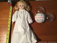 Full view of the porcelain doll lying next to a tape measure showing approximate length and the signed baseball with its protective dome.
