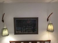 Two teak wall-mounted lamps with fabric shades illuminated on either side of a framed picture above a bed headboard.