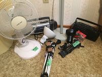 Photo showing white Airworks brand 3-speed fan, a portable Pulsar AM/FM cassette radio, black Sylvania AM radio, and two black and chrome extendable desk lamps with clamps on carpeted floor