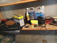 Photo shows an assortment of car care items including jumper cables, boxed Workforce air inflator, tire iron, car mats, washer fluid jugs, small bottles, cloths, and tools arranged on a shelf.