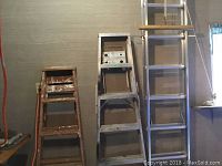 All three ladders standing upright along a textured wall; wooden, aluminum step, and aluminum extension ladder visible.