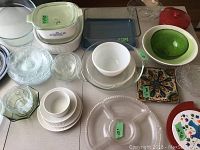 Multiple kitchen glass and ceramic cookware pieces arranged on a table, showing Pyrex items, green ceramic bowl, white Brie pot, decorative trivets, and stacked bowls and plates.