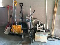 Wide view of full lot showing snow shovels, brooms, vacuum, and hand dolly against garage wall.