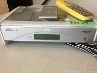 Front view of silver JVC disc player showing digital display, front-loading disc tray, power button, and control panel, with remote control and user manual on top.