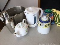 Photo showing Rival electric kettle, two ceramic teapots, martini shaker, metal bucket, and two knitted tea cozies